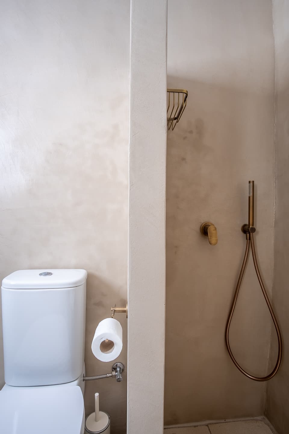 A walk-in shower recess at Casa di Lavalle finished in pale polished plaster, fitted with a brushed brass mixer, handheld shower head, and wall-mounted caddy.