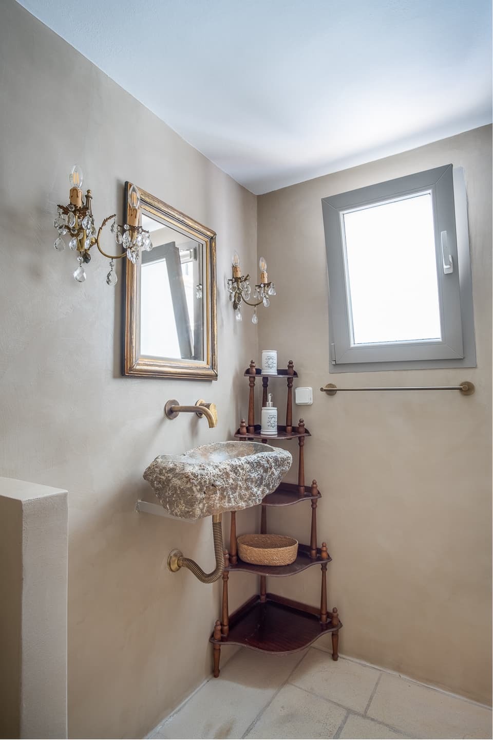 A hand-carved natural stone sink at Casa di Lavalle beneath a gilt-framed mirror and crystal wall sconces, beside a tiered antique corner shelf.